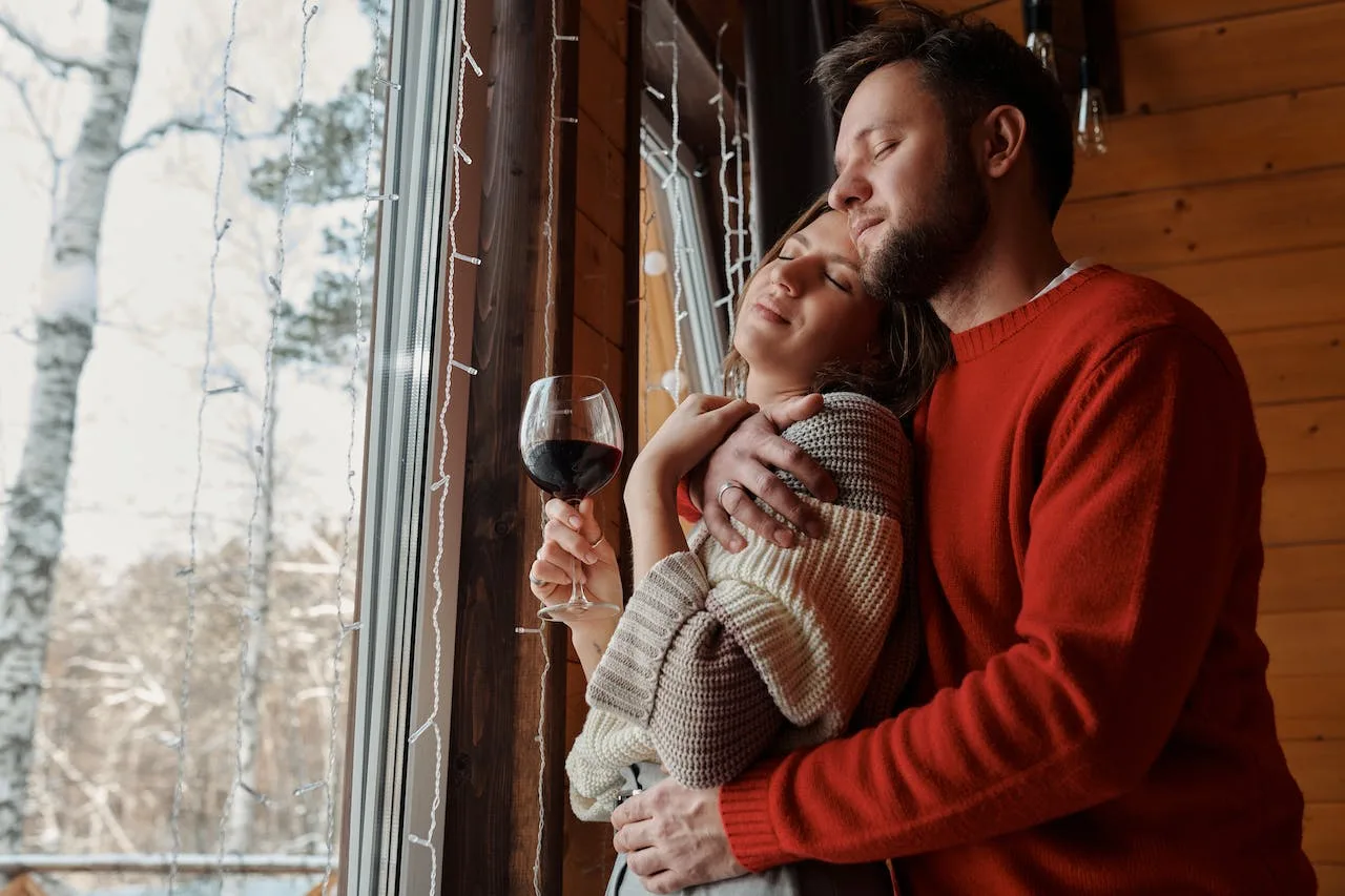 zagrljeni zaljubljeni par sa čašom vina pored prozora